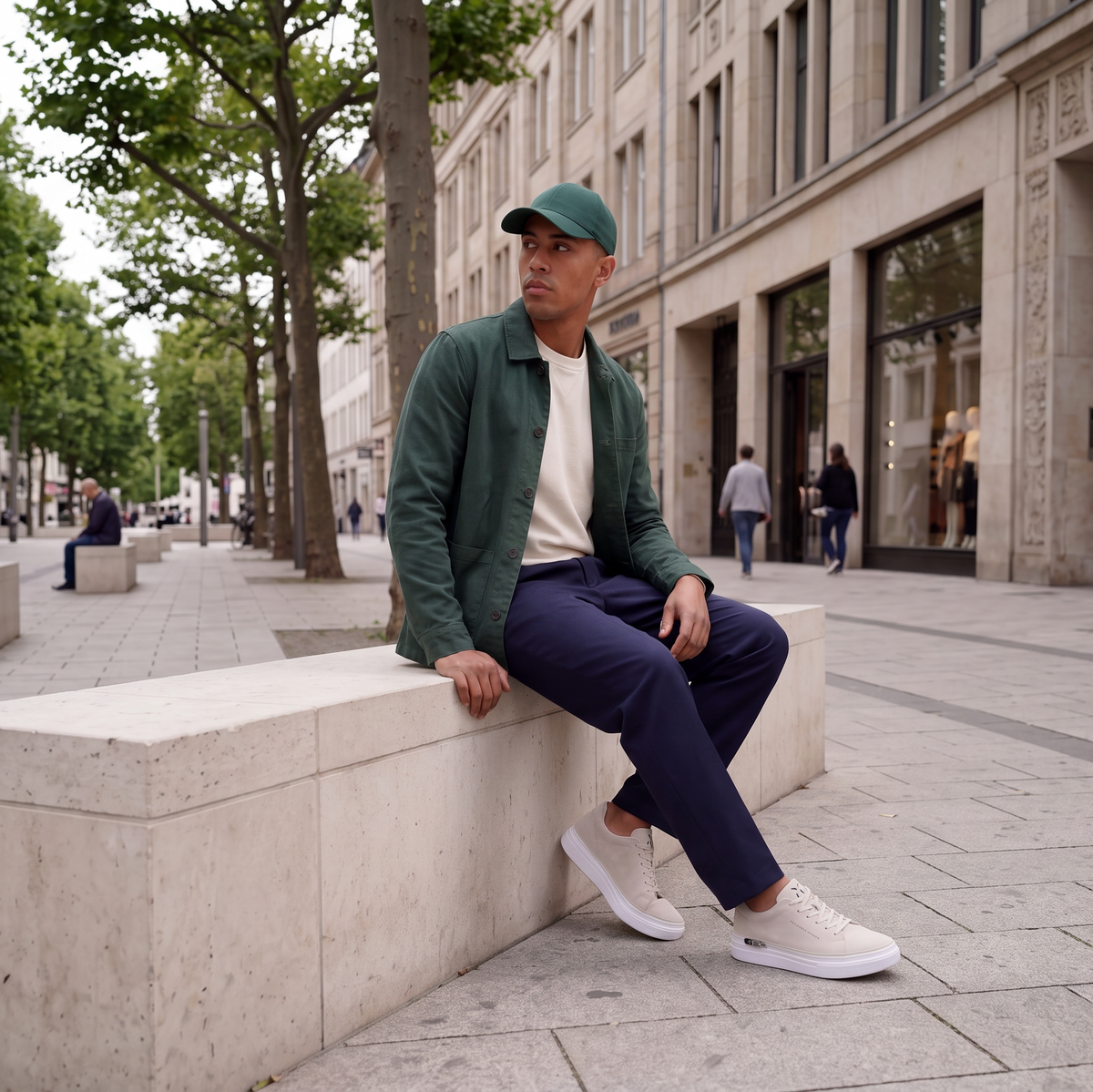 Mann in dunkelgrüner Jacke und dunkelblauer Hose sitzt in einer urbanen Einkaufsstraße und trägt modulare 24couture Horizon Sneaker in Sand auf einer weißen Perpetual Sohle mit Lauffläche in Dune, einem warmen Sandbeige.