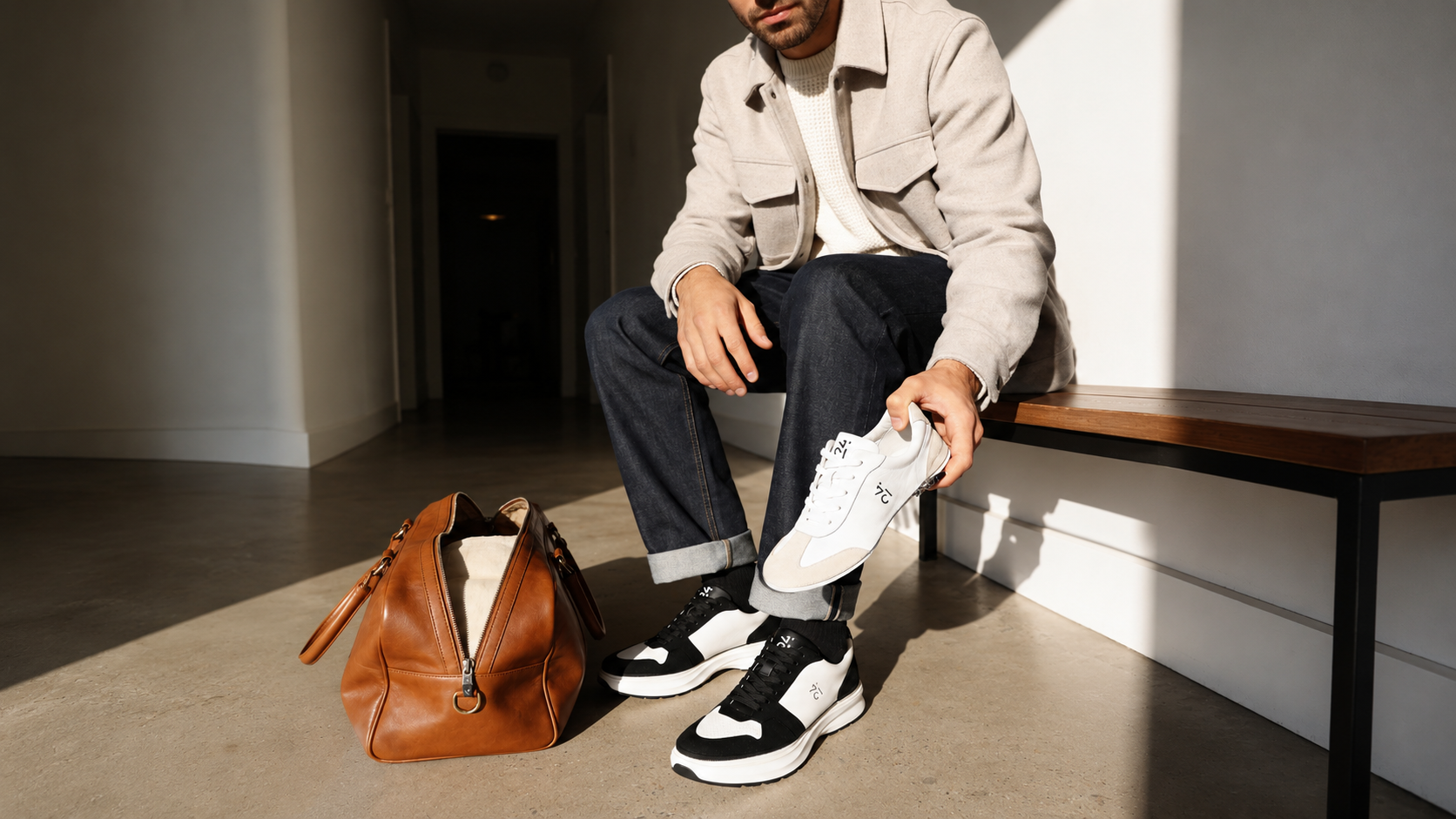 Mann sitzt neben einer braunen Reisetasche und hält ein wechselbares 24couture Adore Upper in White und Sand. Er trägt Ascend Sneaker in White und Black auf der Impulse Sohle in White und Black und demonstriert das modulare Wechselsystem.